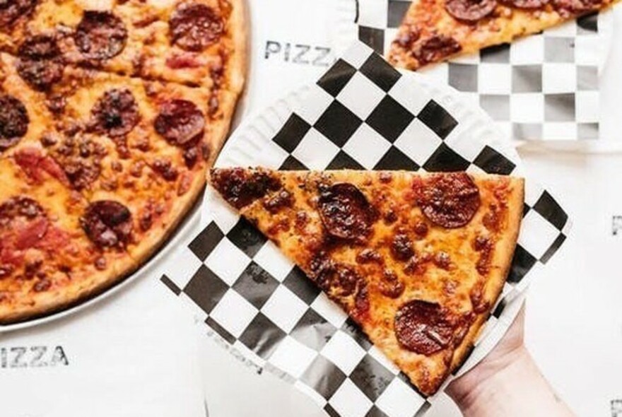 A hand holding a slice of pepperoni pizza on a black and white checked serviette, next to a pizza.