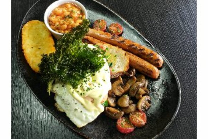 A full English breakfast plate with poached egg, mushrooms, sausages, tomatoes baked beans and a hash brown. 