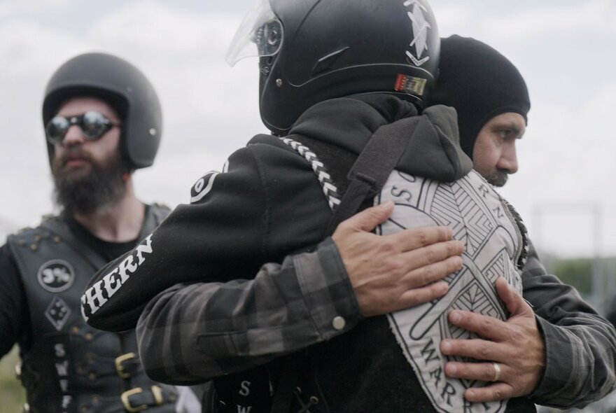 Two members of an Indigenous motorcycle gang embracing, with a third in the background.