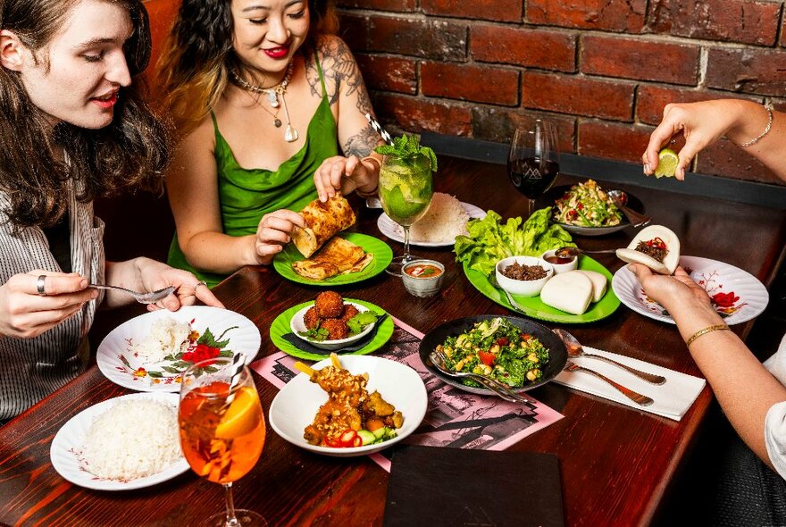 Three women seated at a wooden table with a selected of Thai-style dishes and cocktails.