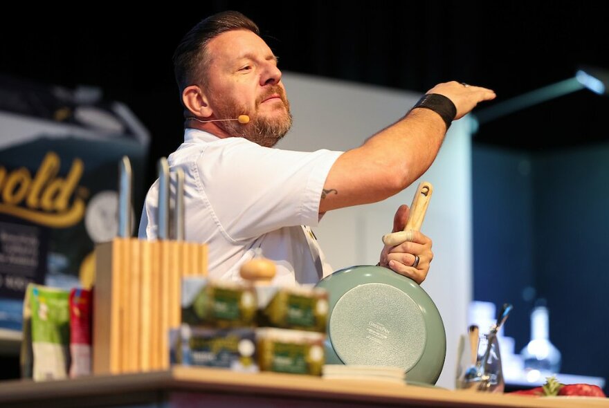 Chef Mani Fidel on stage during a cooking demonstration holding a frying pan and holding up his arm. 
