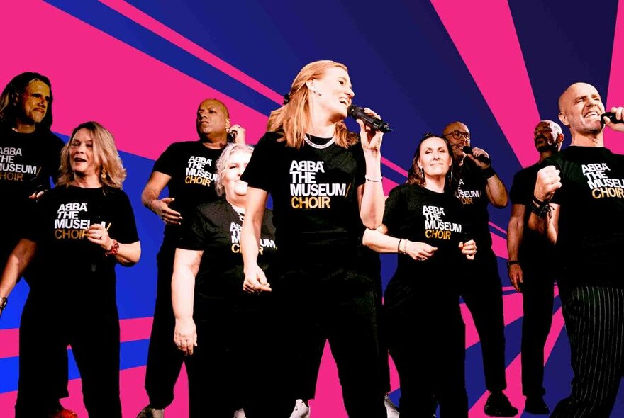 Singing group wearing black T-shirts with ABBA the Choir slogans, holding microphones and dancing against a blue and pink striped background.