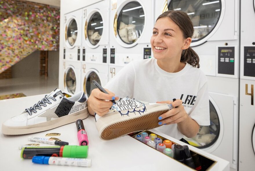 A smiling woman drawing on a pair of sneakers with a texta, in front of a row of washing machines 