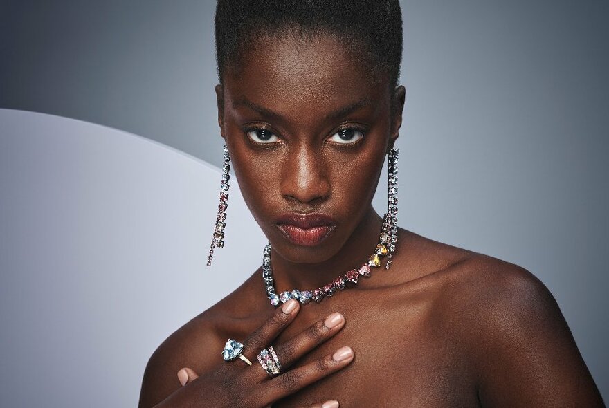Woman from chest up, staring intensely at camera, wearing dangly jewelled earrings, chain and rings.