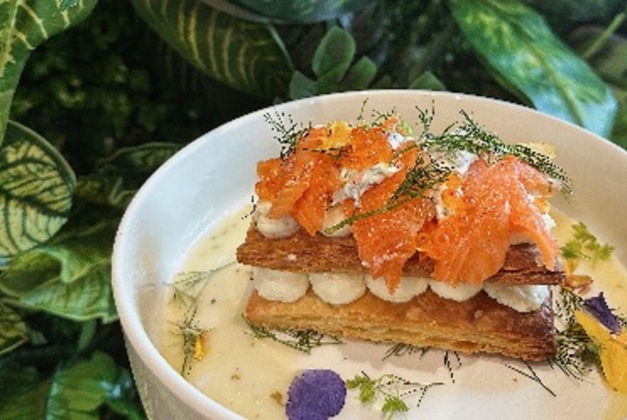 Pastry with salmon and dill on top, in white bowl, greenery behind.