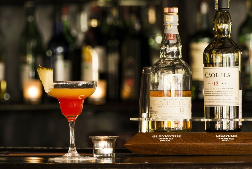 A red and orange cocktail on the counter of the bar, together with two bottles of alcohol.
