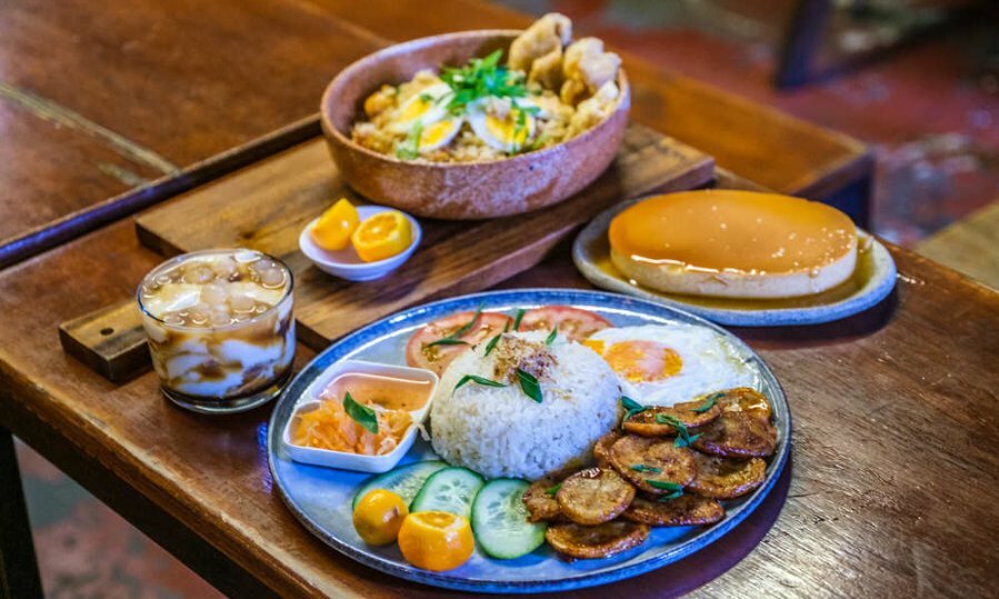 A plate of rice and meat with pickles, various Asian desserts and other dishes on a wooden table in a cafe.