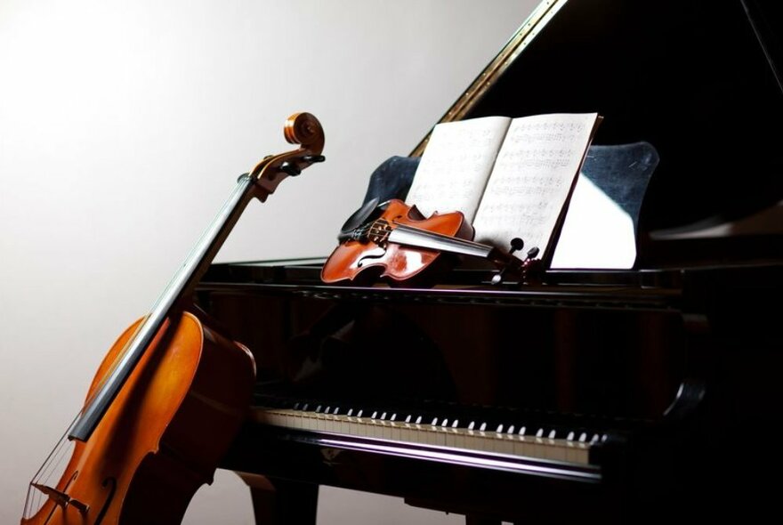 A cello resting on an open grand piano's keyboard, with a violin lying next to sheet music.