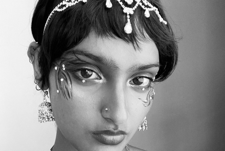 Black and white image of a woman's face with artistic designs around her eyes and a beaded hair decoration. 