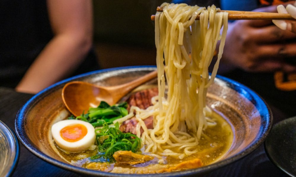 Bowl of ramen with boiled egg, green vegetables and pork. Person holding up the noodles with chopsticks.