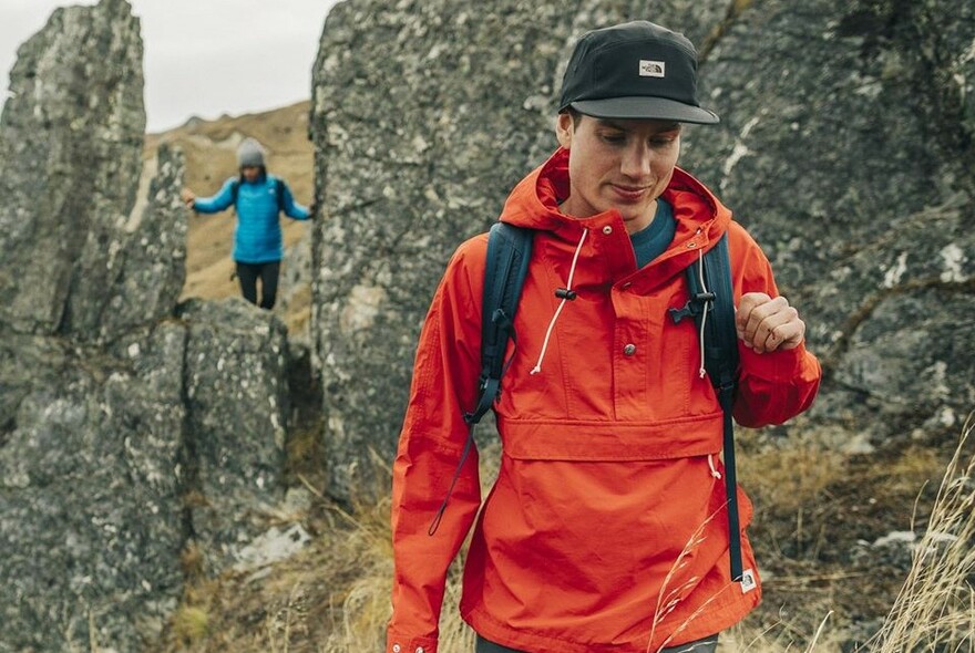 Man wearing an orange hiker's rain jacket and carrying a daypack walking in the rocky outdoors. 