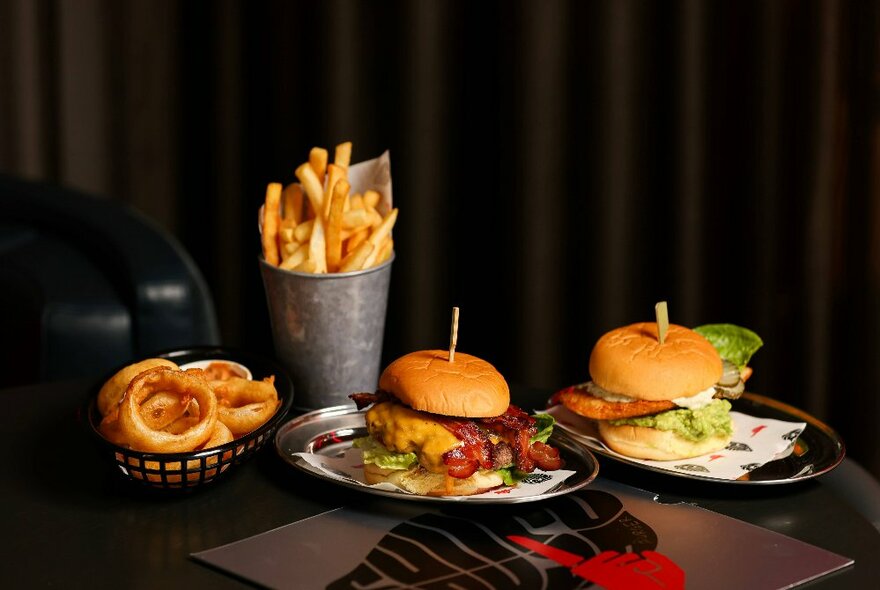 Two gourmet burgers, onion rings and a metal cup of fries displayed on a dark table in front of a dark curtain.