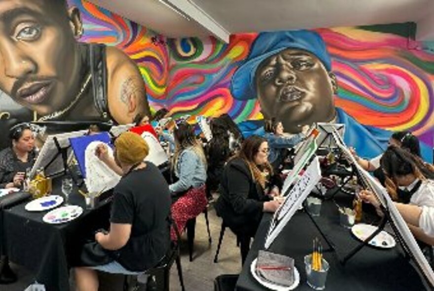 Several rows of people working on canvases in room with large, wall murals of two hip-hop styled men against colourful swirls, at rear and to right.