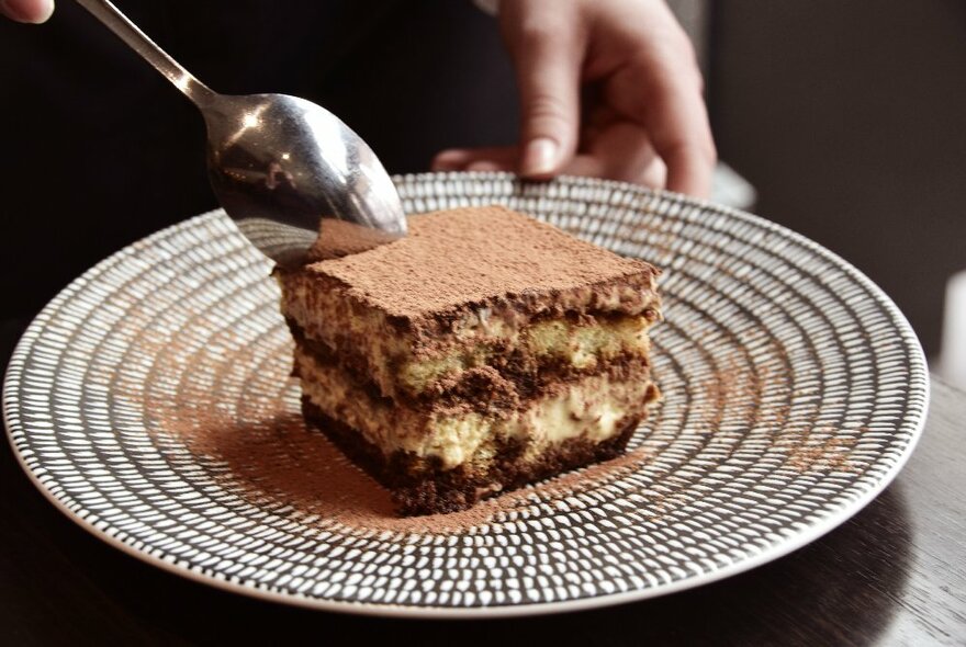 A spoon poised to cut into a large slab of Tiramisu on a decorated plate.