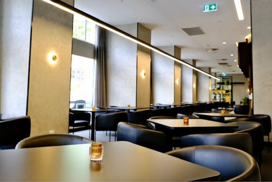 Interior of restaurant showing a row of square tables and black club chairs.