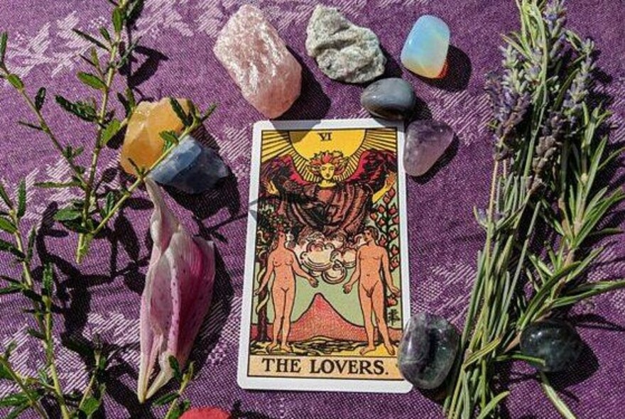 The Lovers tarot card on a purple background surrounded by crystals and herbs.