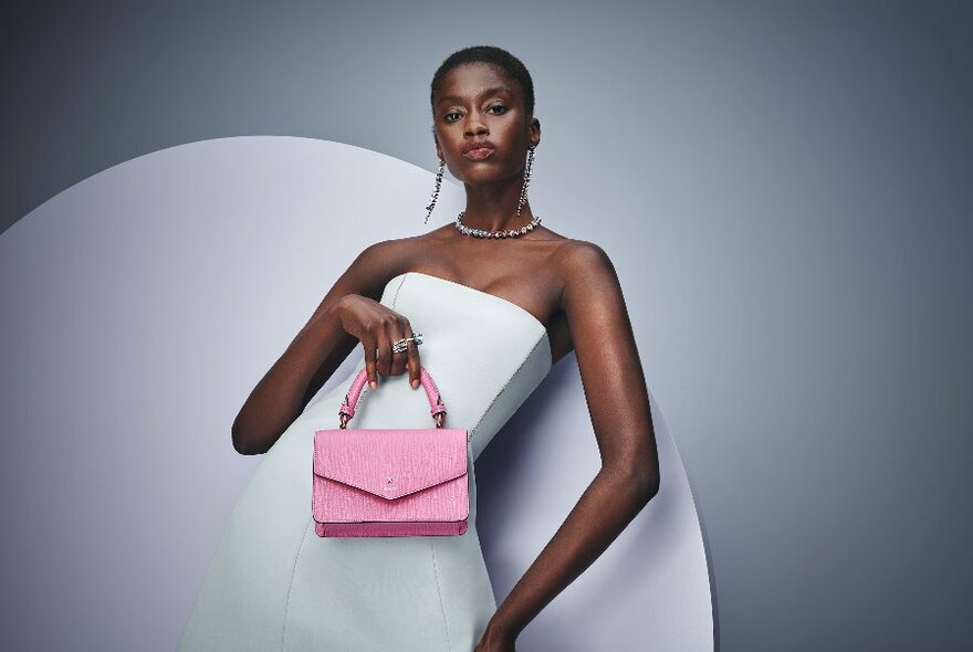 Model in white, strapless dress holding small, light pink bag to waist, against two-toned grey backdrop.