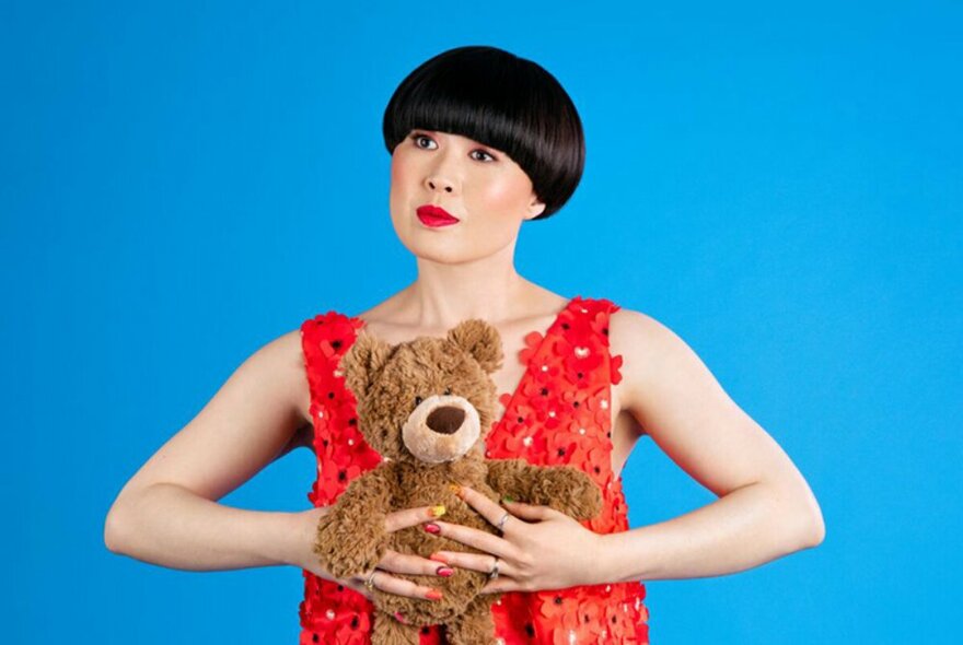 Comedian Atsuko Okatsuka with a bowl haircut, wearing a red dress and holding a teddy bear.