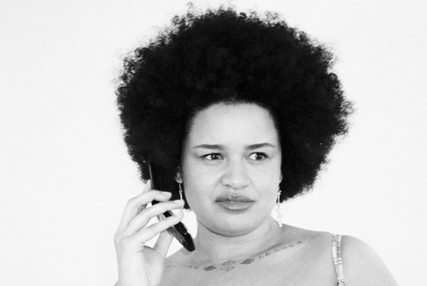 Black and white image of a woman with tight curly hair looking quizzical holding up her phone. 