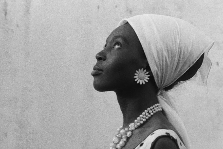 A black and white profile portrait of an African woman wearing a white headscarf, flower earrings and a pearl necklace.