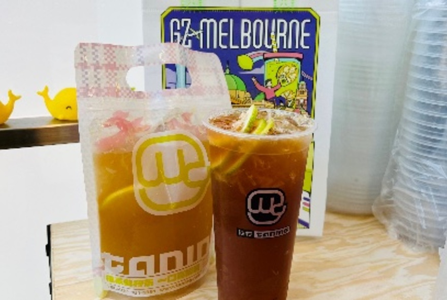 A plastic takeaway cup of iced tea and a sealed plastic takeaway bag of iced tea, resting on a counter inside a shop.