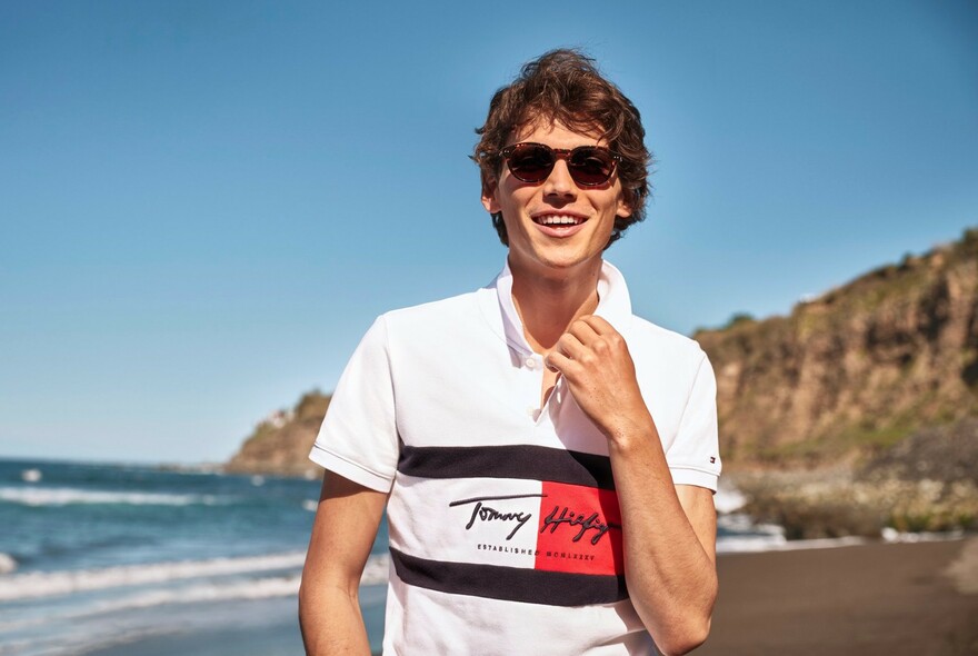 Male model on a beach wearing Hilfiger logo polo and sunglasses.