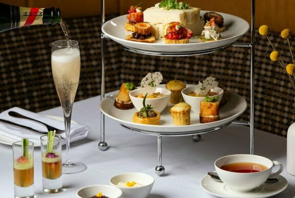 White linen topped table with a tiered stand of savoury and sweet high tea treats, tea in china cups and champagne being poured from a bottle into a glass.