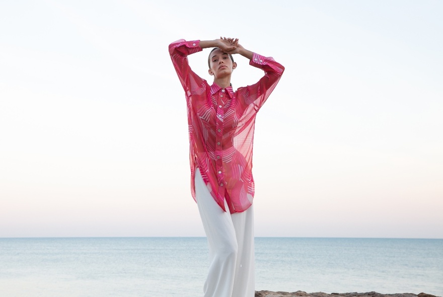 Female model wearing a red chiffon long shirt over white trousers, posing at a beach with the sea behind her.