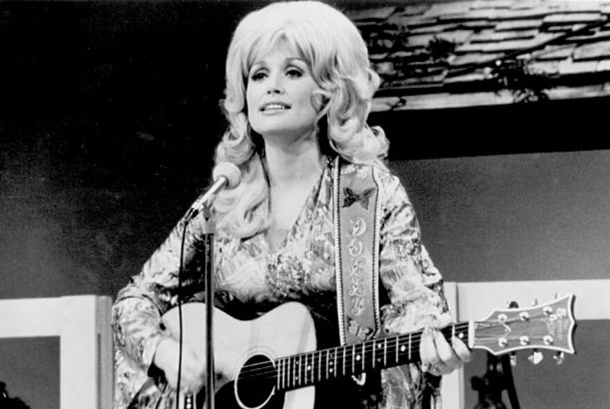 An old black and white photo of Dolly Parton with big hair playing an acoustic guitar.