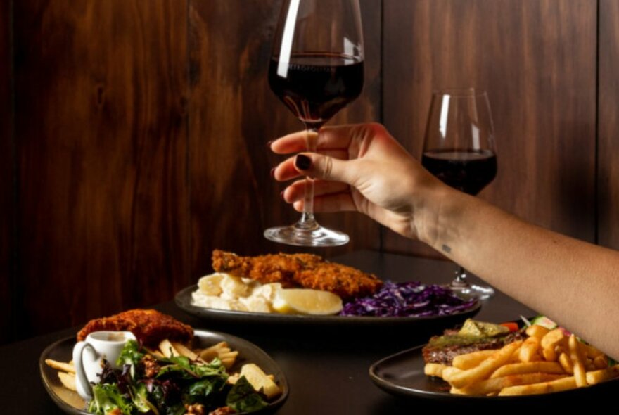 A hand holding a glass of red wine over a table set with plates of food including chips and salad.