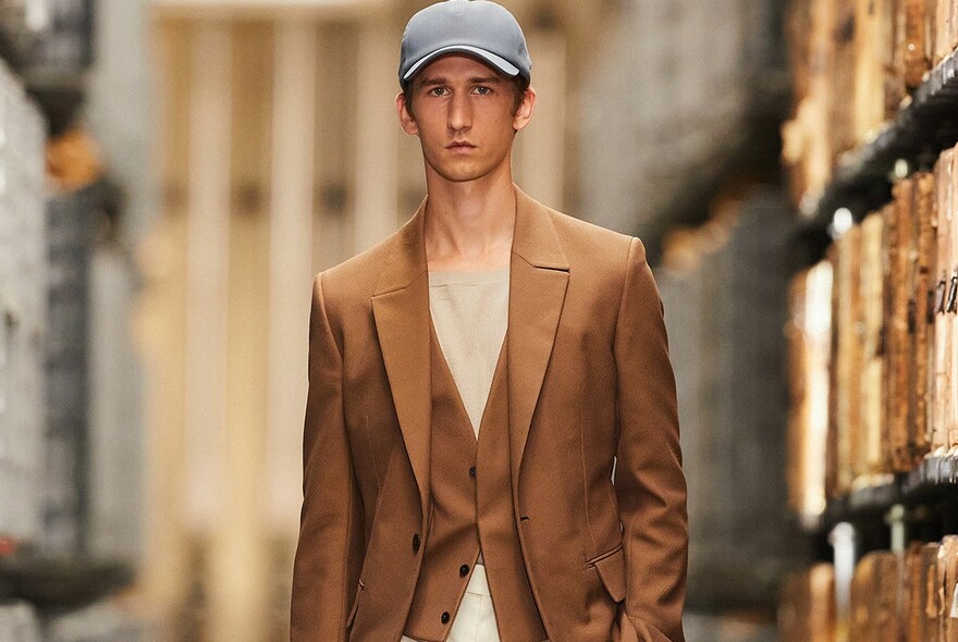 Male model wearing brown suit, beige T-shirt and light blue cap.