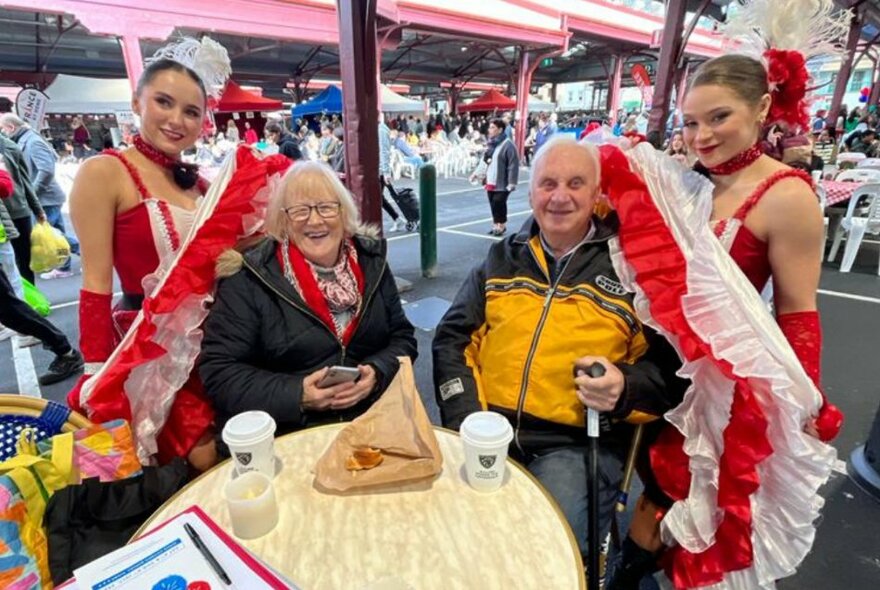An older couple seated at a cafe table with two French cabaret dancers in red and white posing behind.