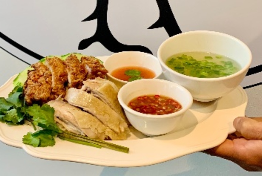 Pair of hands holding a large plate of poached chicken over rice and salad with bowls of dipping sauces of the side.
