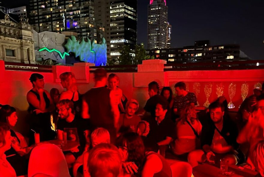 Rooftop outdoor bar area with people sitting around drinking and talking, seeped in red light, with a night time view of the Melbourne city skyline.