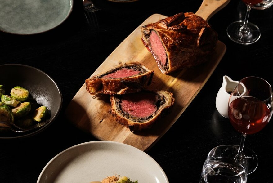A wooden serving board with King Island Beef Wellington surrounded by accompanying dishes and wine.