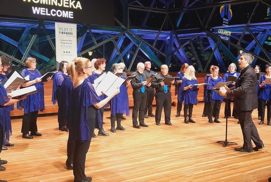 The Choir singing in a venue with wooden floor and purple scaffolding, led by a conductor.