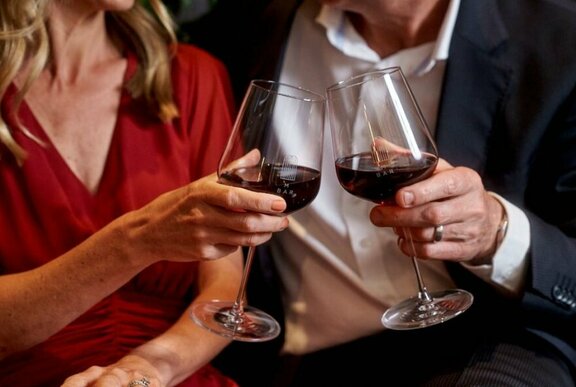 Male and female clinking glasses of red wine, wearing relaxed semiformal dress and open-necked shirt with suit.