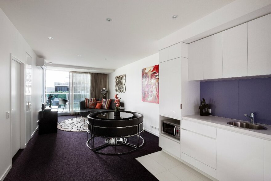Sebel Docklands hotel apartment with purple carpet and white walls, open-plan living and kitchen leading to balcony.