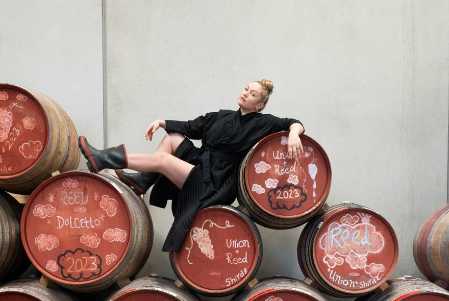 A young winemaker reclining on barrels of wine, wearing a dress and gumboots.