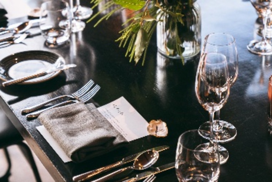 A dark timber table set for dinner, with multiple wine glasses, gold cutlery and a linen napkin.