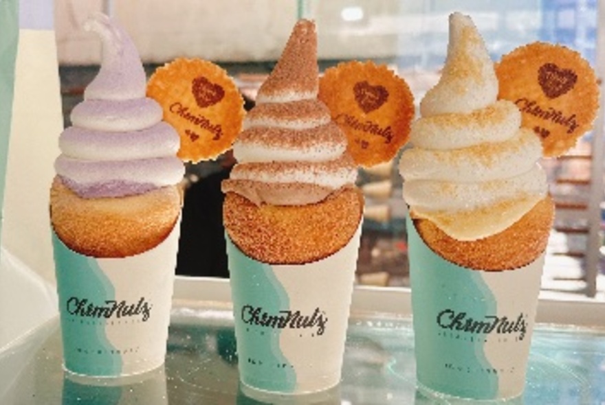 Three chimney cakes topped with different flavoured ice creams standing in a row in paper cones. 